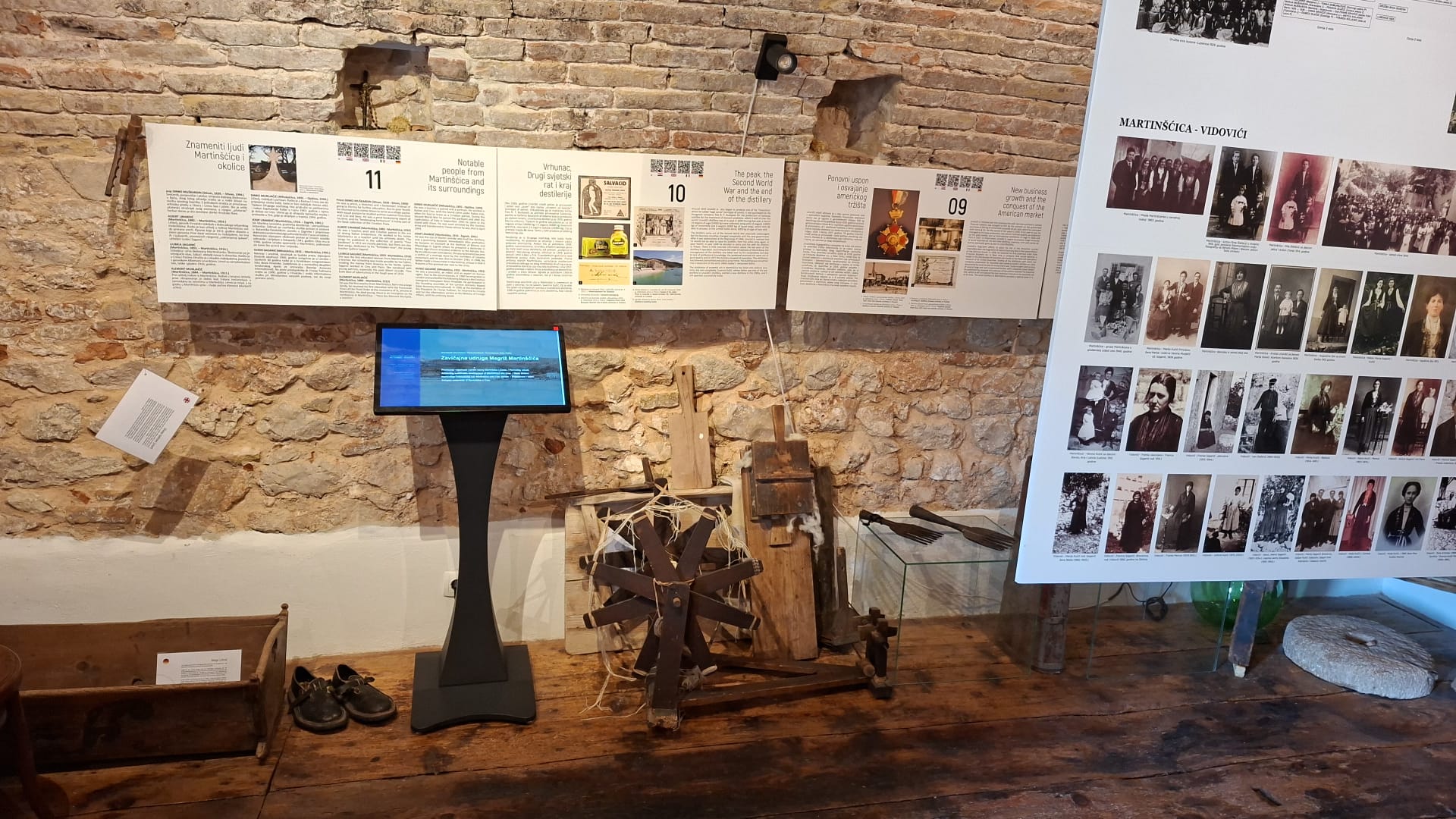 Exhibition display with informational posters, digital screen, and historical artifacts against a stone wall.